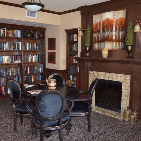 A cozy interior room featuring a round wooden table surrounded by four upholstered chairs with dark fabric. Behind the table is a built-in wooden bookshelf filled with books. To the right, there is a fireplace with a granite surround and a wooden mantel decorated with two topiary plants and a ceramic vase. Above the mantel hangs a painting of trees with autumn-colored leaves. The room has beige walls, dark wood trim, and a patterned carpet.