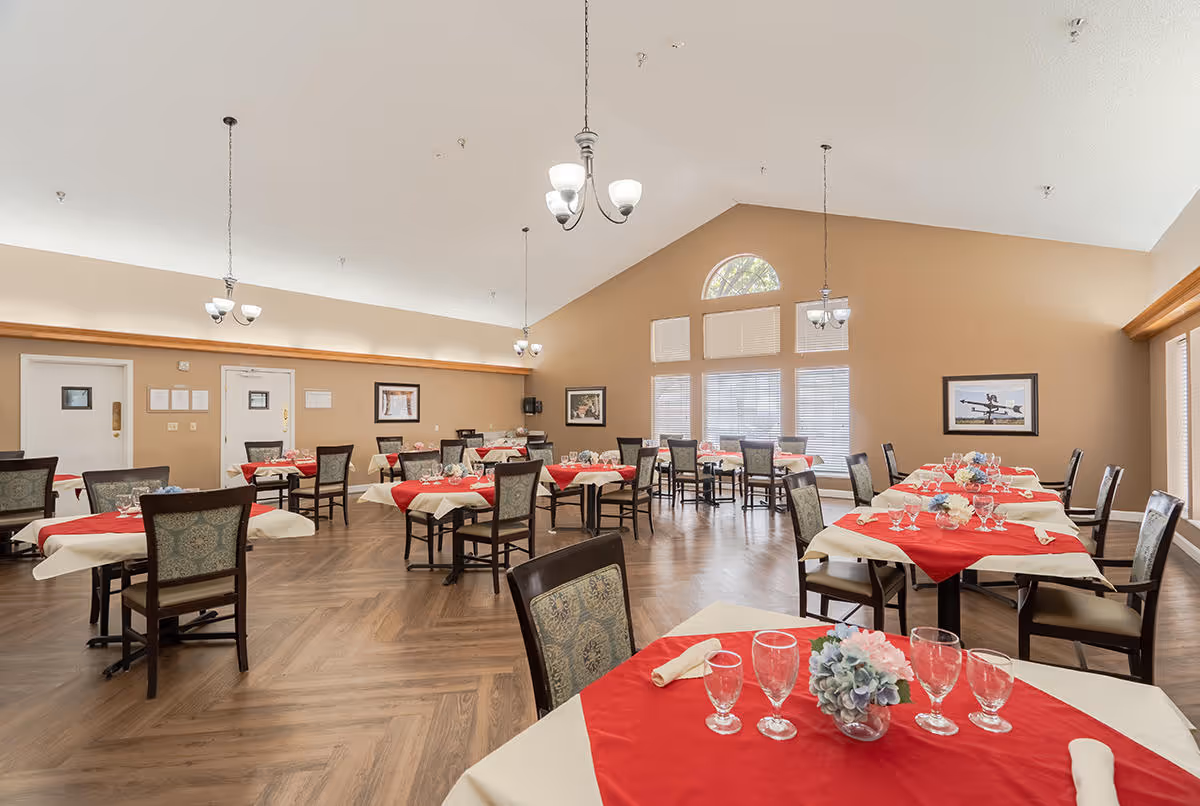 A spacious dining room with multiple tables covered in white tablecloths and red table runners. Each table is set with glassware, napkins, and small floral centerpieces. The room has high vaulted ceilings with hanging light fixtures, large windows with blinds, and framed artwork on the beige walls. The floor is wood with a herringbone pattern.