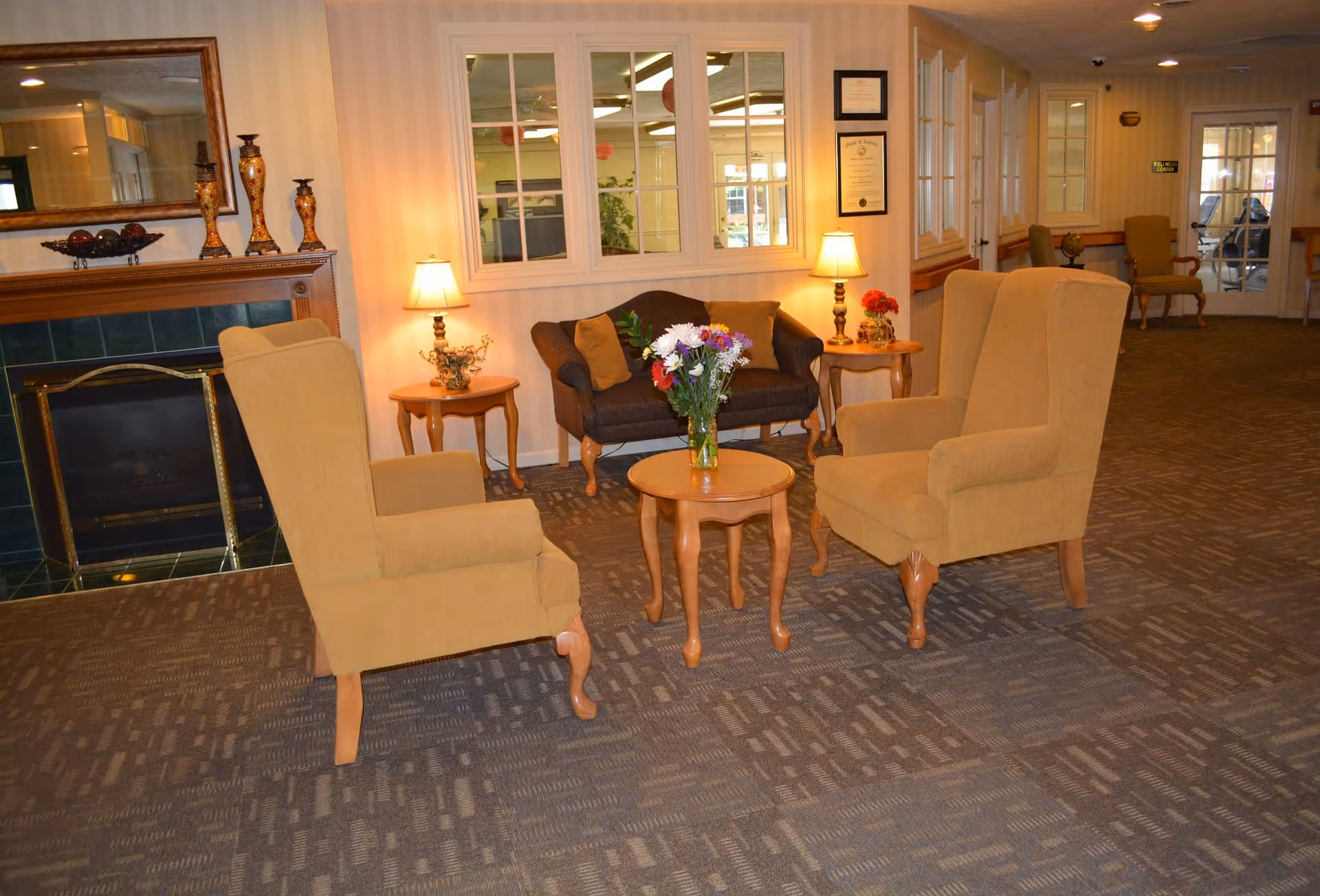 A cozy sitting area in a senior living facility with two beige armchairs and a small round wooden table with a vase of flowers in the center. Behind the table is a dark brown loveseat with two cushions, flanked by two side tables each with a lamp. A fireplace with decorative items and a large mirror above it is visible on the left side. The room has carpeted flooring and light-colored walls with windows and framed certificates.
