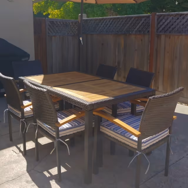 Outdoor patio area with a rectangular wooden table surrounded by six wicker chairs with striped cushions, set on a concrete surface next to a wooden fence and a closed umbrella.