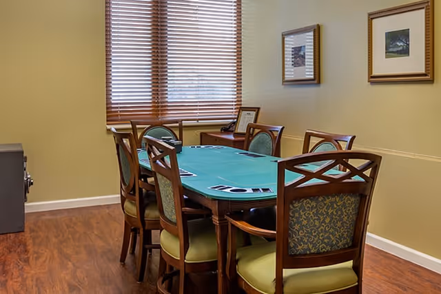 Game room with a green felt poker table surrounded by wooden chairs, a window with blinds, and framed artwork on the wall.