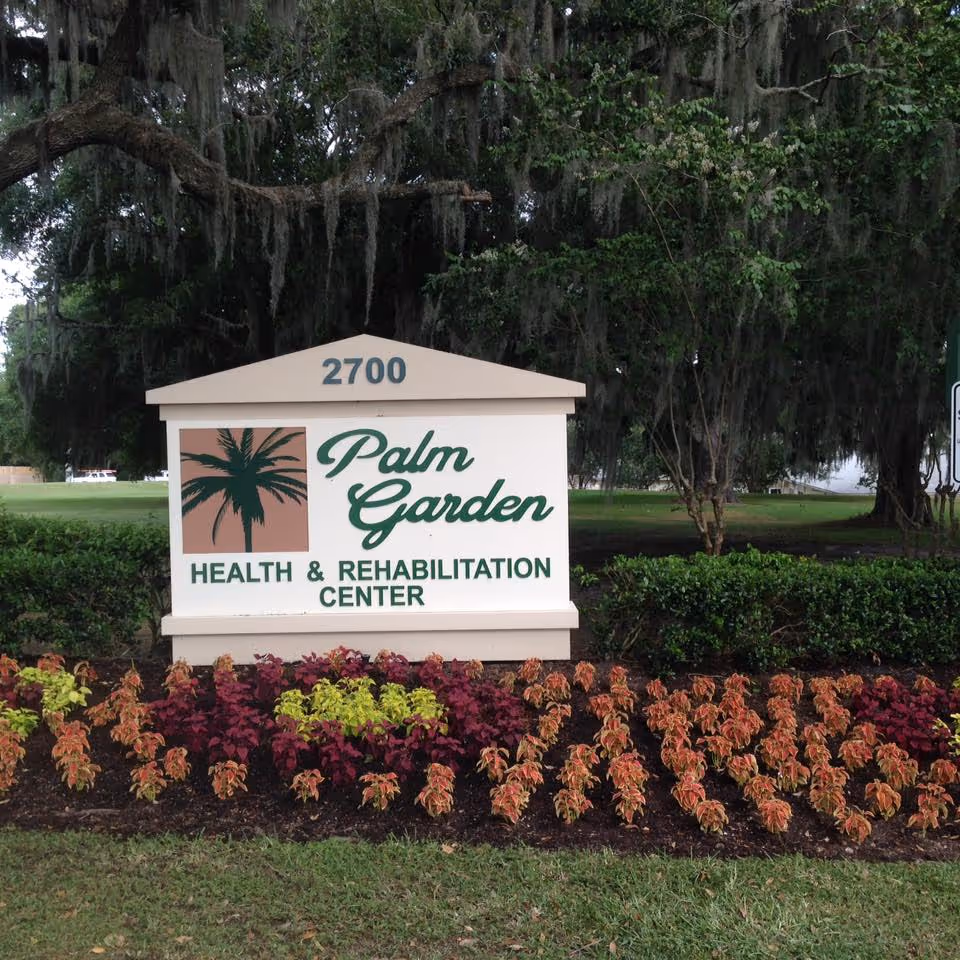 Entrance sign reading "Palm Garden Health & Rehabilitation Center" with the number 2700, set among landscaped flowers and large oak trees.