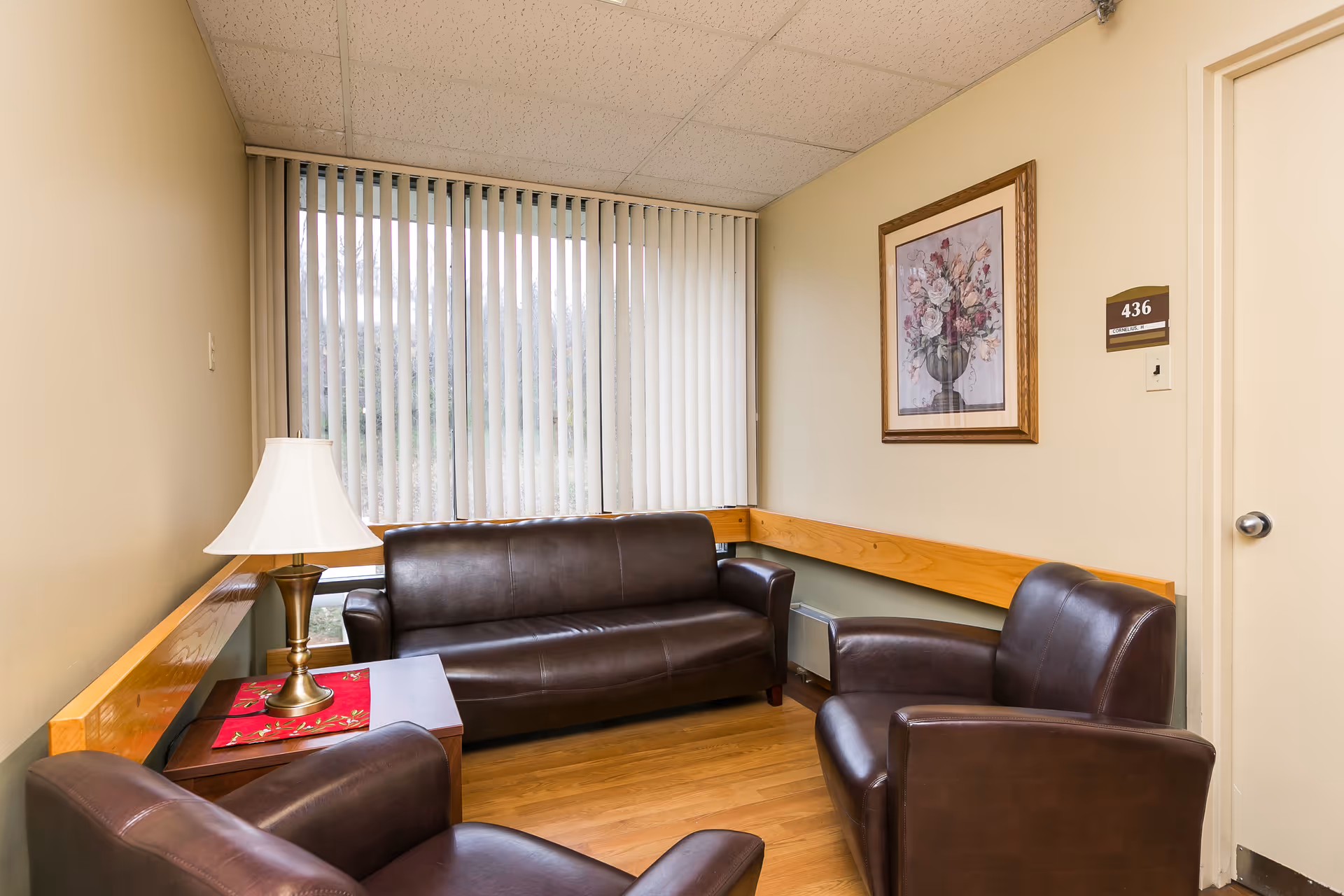 A small sitting area in a nursing facility with a brown leather couch, two matching brown leather armchairs, a wooden side table with a lamp, vertical blinds covering a window, a framed floral picture on the wall, and a door labeled 436.