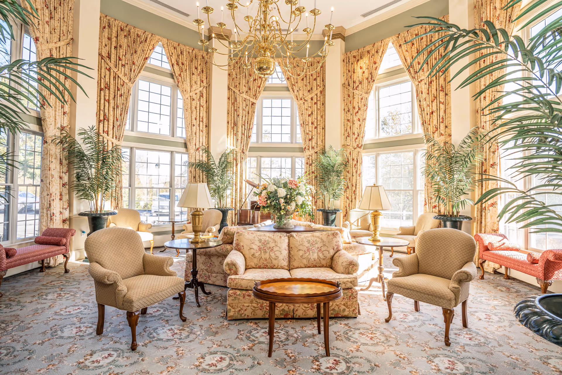 A bright and elegant living room with large windows covered by floral curtains, multiple upholstered armchairs and sofas arranged around wooden tables, potted plants, and a chandelier hanging from the ceiling.