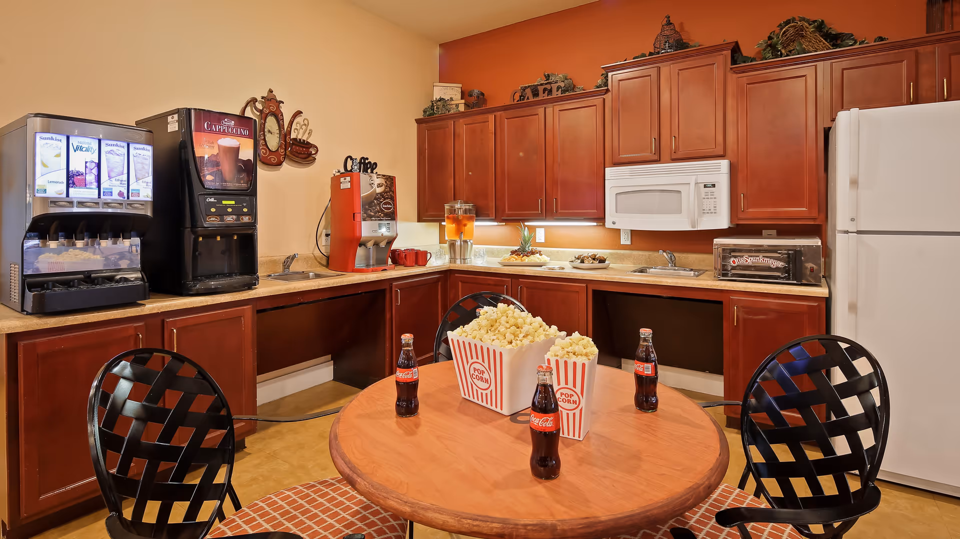 A kitchen area with wooden cabinets, a white microwave, and a white refrigerator. On the countertop, there is a cappuccino machine, a juice dispenser, and a coffee maker. In the foreground, a round wooden table with three black chairs has three bottles of Coca-Cola and two containers of popcorn on it.