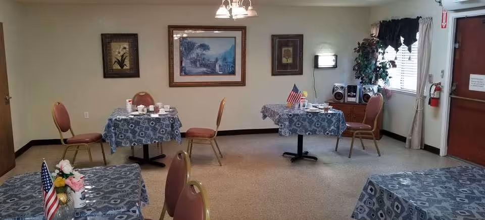 Small communal dining room with several round tables covered in patterned tablecloths, chairs, framed wall art, and an American flag on a table.