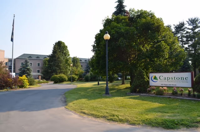 Exterior view of Capstone Center for Rehabilitation and Nursing showing a paved driveway, green lawn, trees, a flagpole with a flag, and a sign with the facility's name near the entrance.