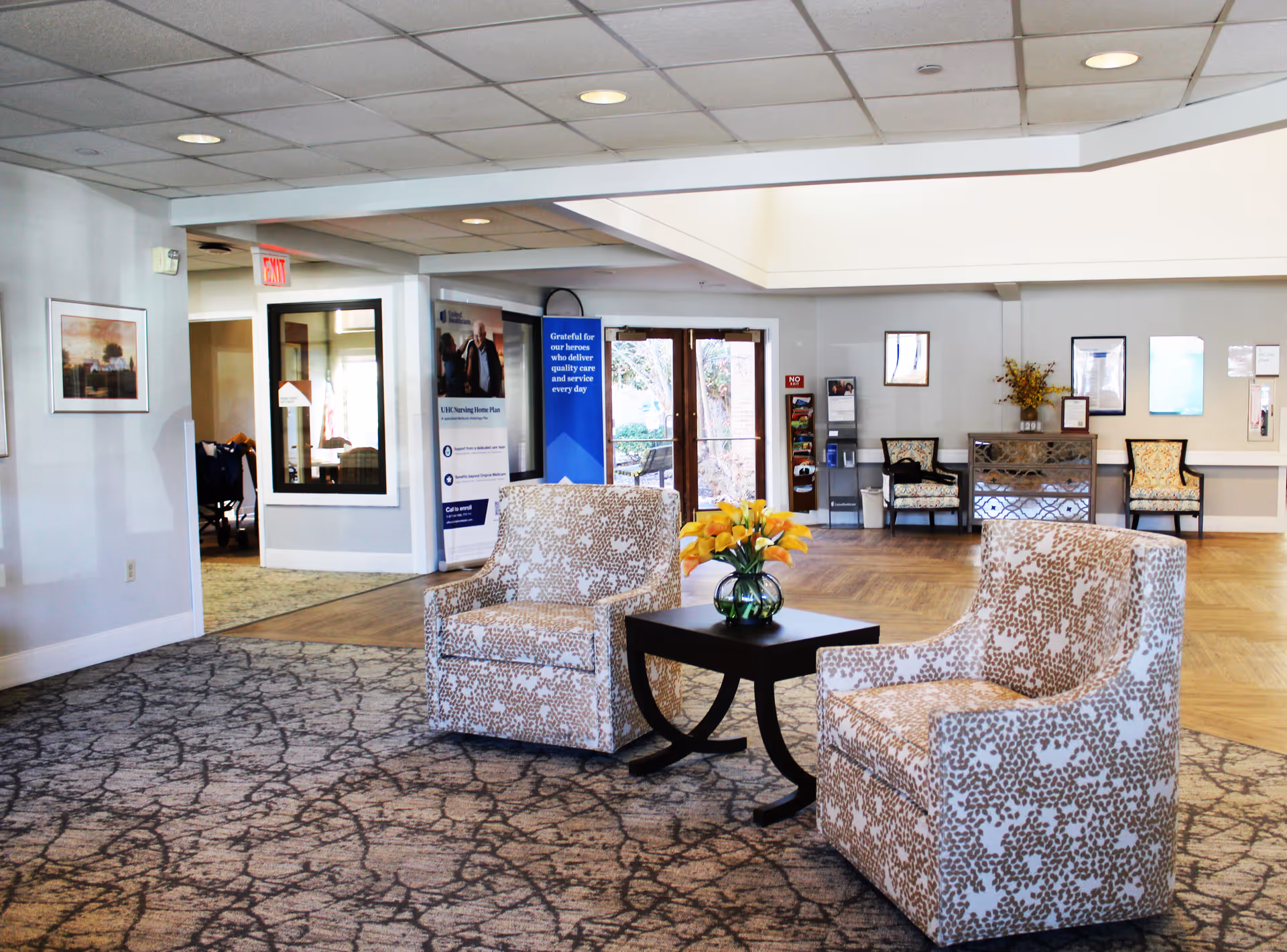 A bright and spacious common area in Sherbrooke Village Living Center featuring two patterned armchairs facing each other with a small black table in between holding a vase of yellow flowers. The room has a mix of carpet and wood flooring, with additional seating and framed pictures on the walls. Large windows and glass doors allow natural light to enter the space.