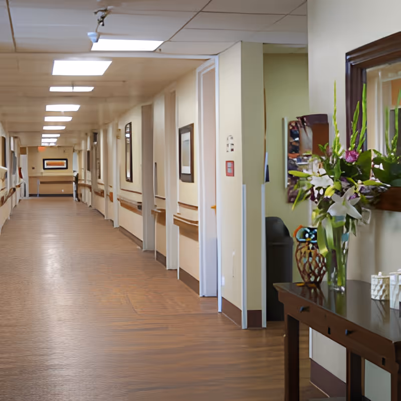 A long, well-lit hallway in a care center with wooden handrails along the walls and framed pictures hanging. On the right side, there is a wooden table with a vase of fresh flowers and decorative items, next to a mirror.