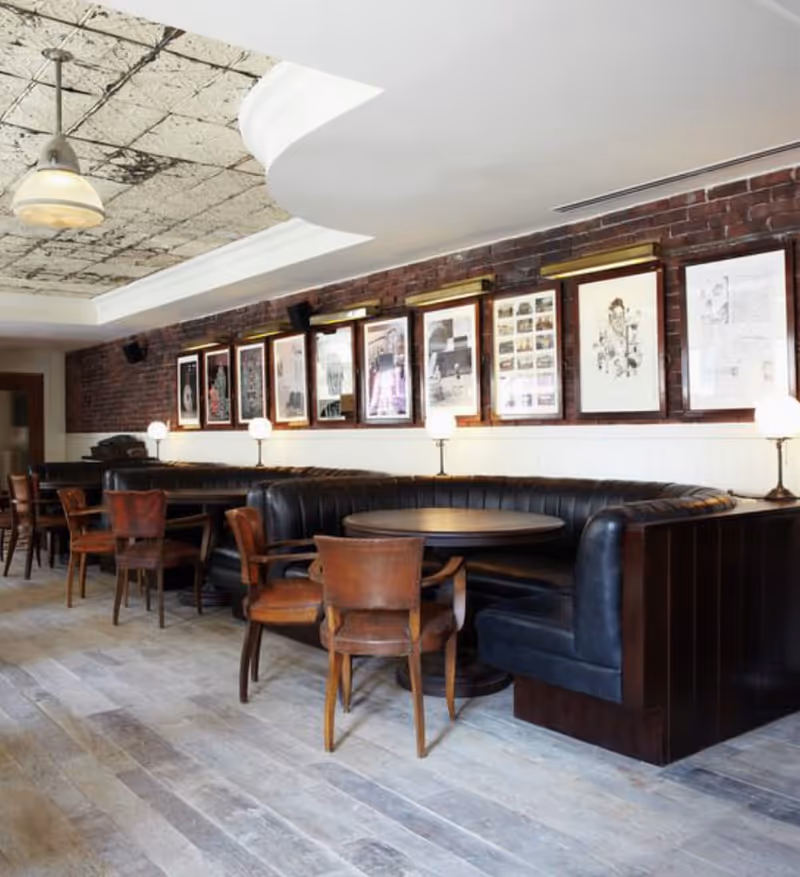 Cozy dining area with curved black leather booths, wooden tables and chairs along an exposed brick wall lined with framed artwork.
