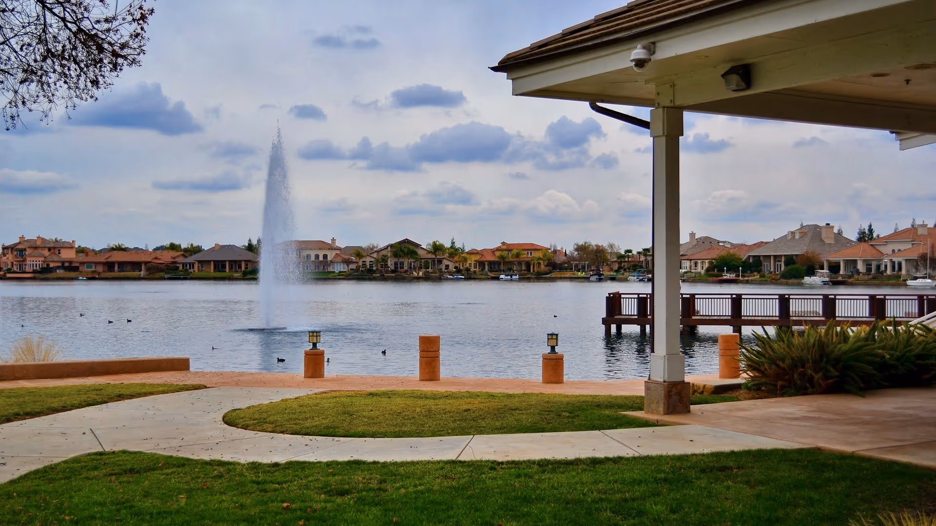 View of a lakeside area with a water fountain in the middle of the lake, surrounded by houses. In the foreground, there is a covered patio area with a walkway, green grass, and some bushes.