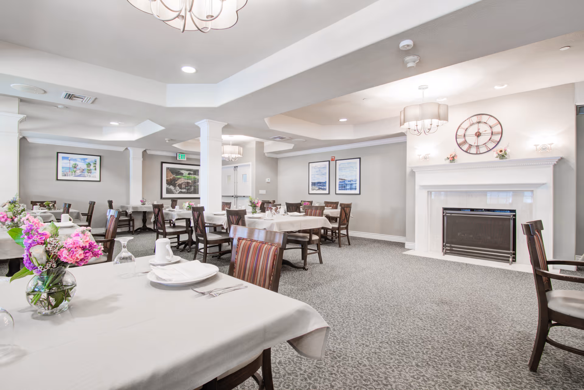 A bright and spacious dining room with multiple tables covered in white tablecloths, set with plates, cups, and silverware. There are wooden chairs around the tables, floral centerpieces, a white fireplace with a large clock above it, and framed artwork on the walls. The room has a carpeted floor and ceiling lights.