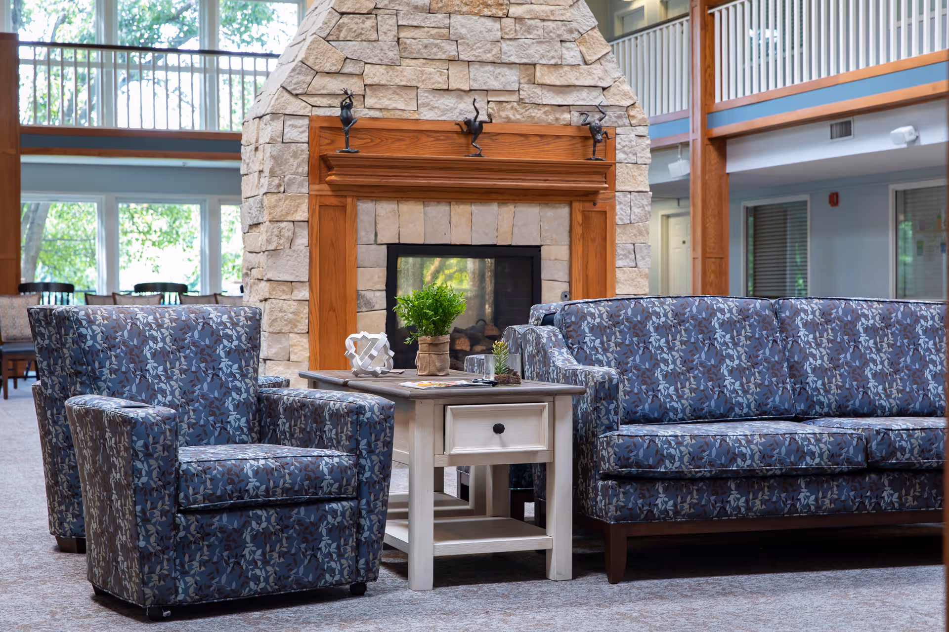 A cozy seating area in a senior living facility featuring a stone fireplace with a wooden mantel, a blue patterned armchair, a matching blue patterned sofa, and a white side table with decorative items and a small potted plant. Large windows and a balcony railing are visible in the background.