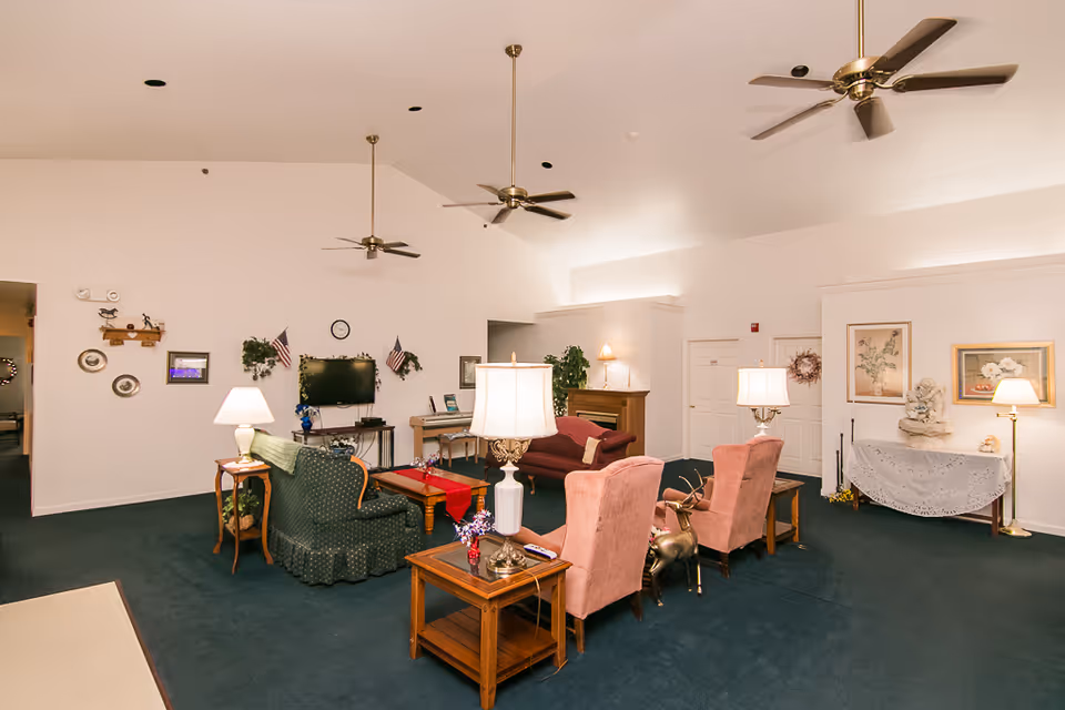 A spacious living room area in Serenity Suites Assisted Living with green carpet, multiple ceiling fans, and various seating options including green and pink upholstered chairs and sofas. The room is decorated with table lamps, framed pictures, American flags, a TV mounted on the wall, and a piano. There is also a small table covered with a lace cloth and decorative items on it.