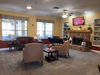 A cozy living room area with beige walls and carpeted floor. The room features two armchairs and a small table with a flower vase in the center. There is a fireplace with a TV mounted above it, displaying a red screen. Shelves with decorative items flank the fireplace. Large windows with valances allow natural light into the room. A ceiling fan with lights is mounted on the ceiling.