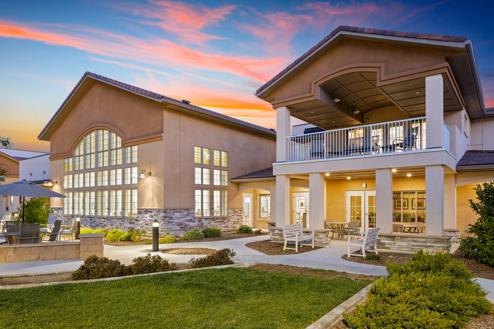 Exterior view of a senior living facility at sunset with well-lit windows, outdoor seating areas including tables, chairs, and benches, surrounded by landscaped greenery and pathways.