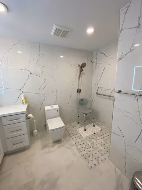 Bright modern bathroom with marble-look tile walls and floor, a toilet and vanity on the left, and a walk-in shower area with a shower chair and hand shower.