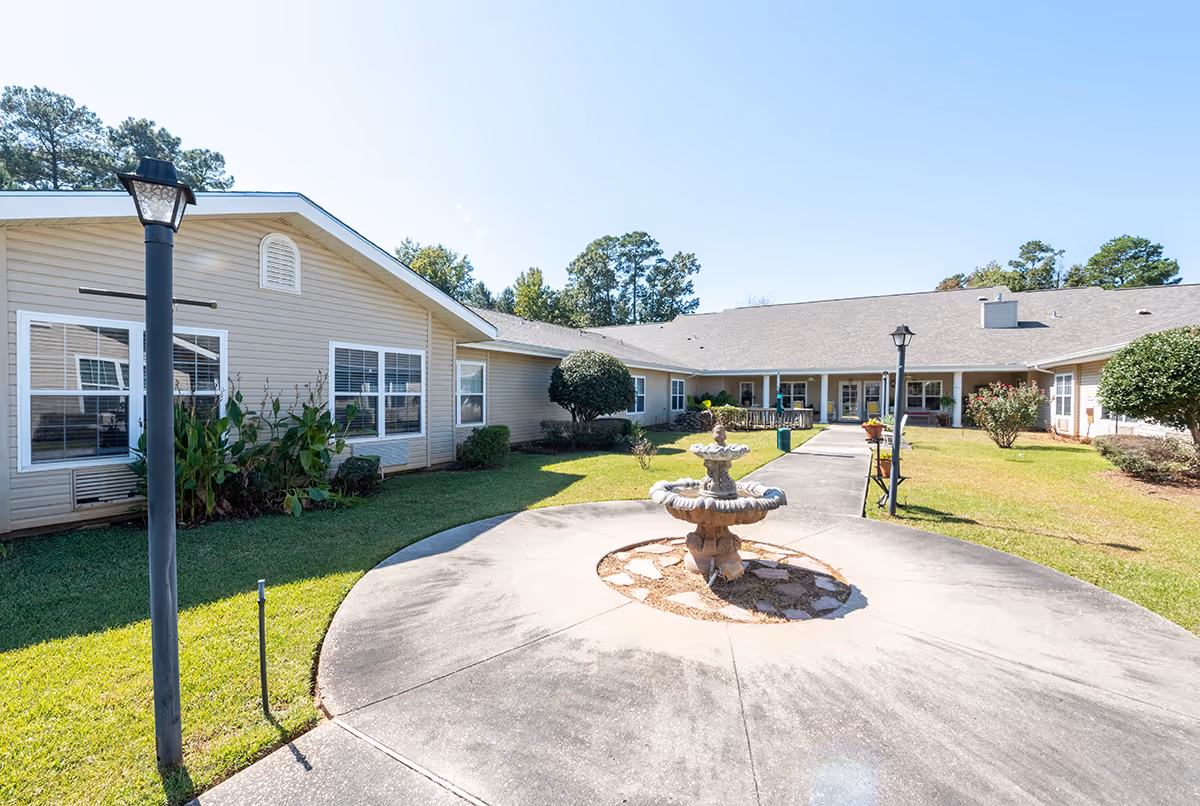 Sunny courtyard of a single-story senior living facility with a central fountain, lamp posts, walkways, and surrounding building wings.