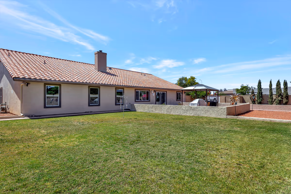 Outdoor view of a single-story building with a tiled roof and several windows. In front of the building is a large grassy lawn, and to the right is a patio area with a gazebo and outdoor grill. The sky is clear with a few wispy clouds.