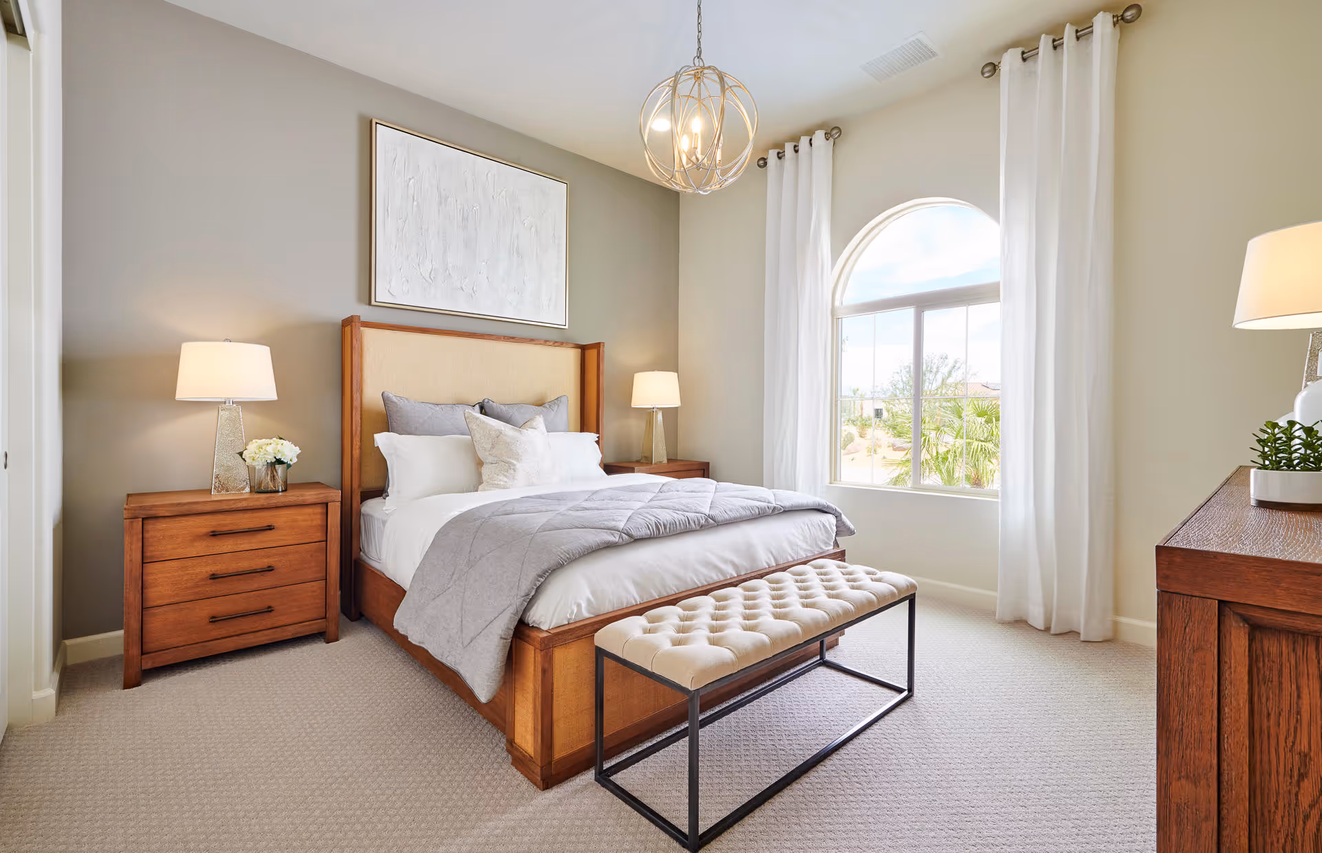 A bright and neatly arranged bedroom featuring a wooden bed with a beige upholstered headboard, white and gray bedding, and multiple pillows. On either side of the bed are matching wooden nightstands with lamps and decorative items. A tufted bench with a metal frame is placed at the foot of the bed. The room has a large arched window with white curtains, beige carpet, and a modern chandelier hanging from the ceiling.