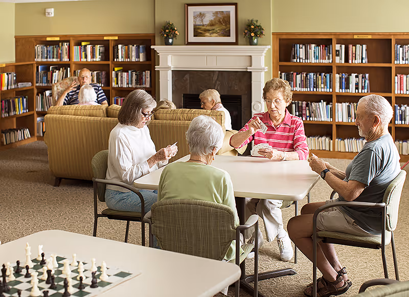 A group of elderly people sitting around a table playing cards in a cozy room with bookshelves and a fireplace in the background. Another table with a chessboard is visible in the foreground.