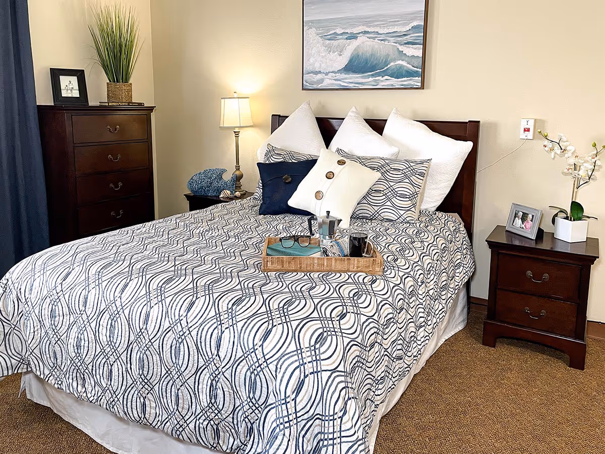 A neatly made bed with a patterned blue and white bedspread and multiple pillows, including decorative ones, in a senior living facility bedroom. There is a wooden nightstand on each side of the bed, one with a lamp and decorative items, and the other with a framed photo and a potted orchid. A wooden dresser with a plant and framed picture is visible to the left, and a painting of ocean waves hangs on the wall above the bed.