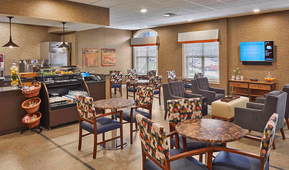 Cozy senior living dining area with round tables, patterned chairs, a small café counter and lounge seating.