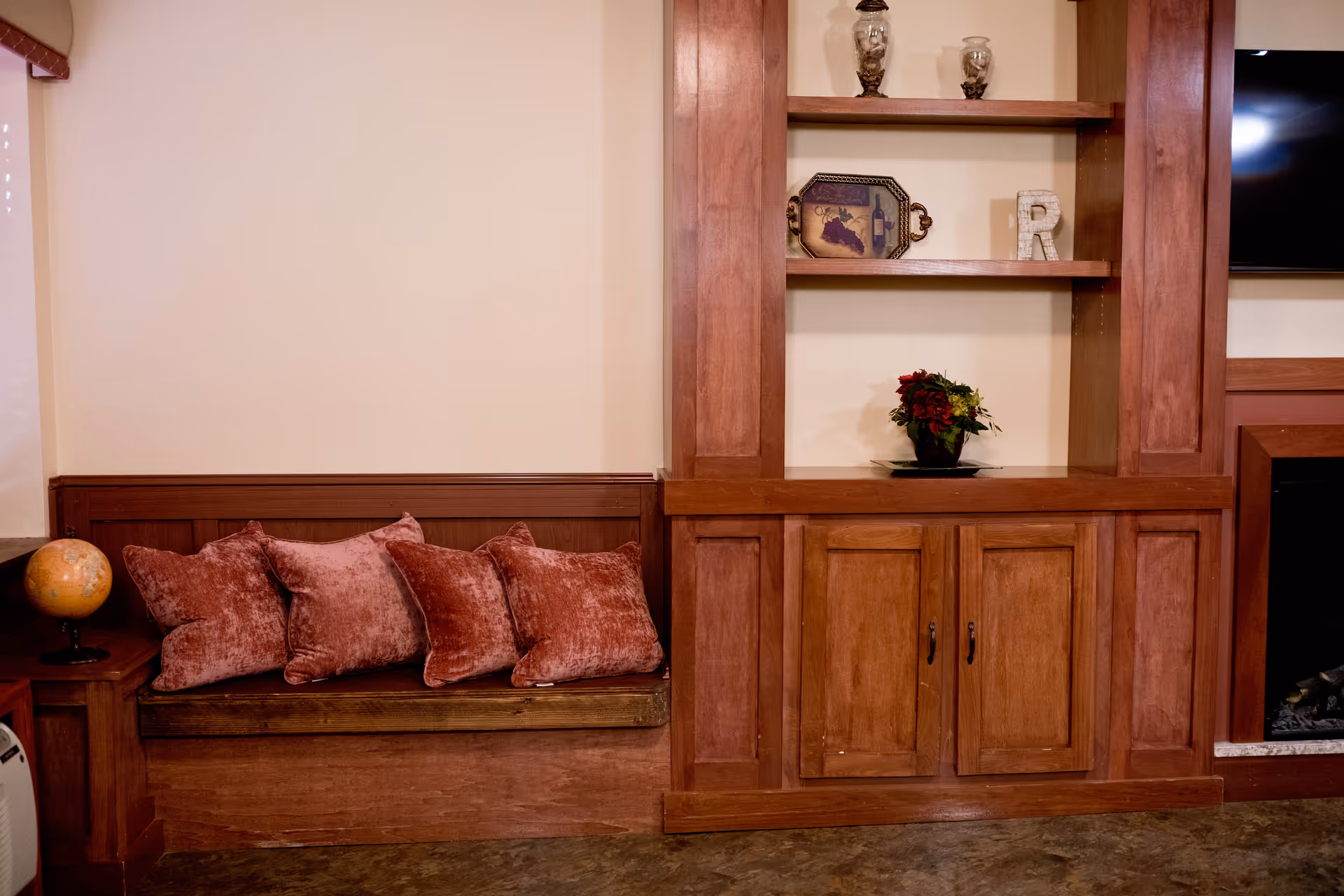 A cozy corner of a living room featuring a built-in wooden bench with five plush red cushions, a wooden cabinet with shelves displaying decorative items including a small globe, a tray, a letter 'R', and a flower arrangement. A flat-screen TV is partially visible on the right side above a fireplace.