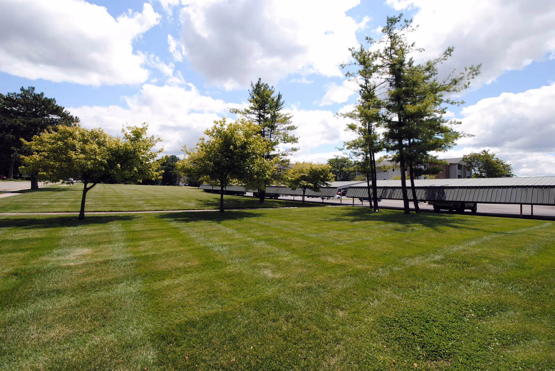 A well-maintained grassy lawn with several small and medium-sized trees under a partly cloudy sky. In the background, there is a covered parking area with cars and a building partially visible behind the trees.