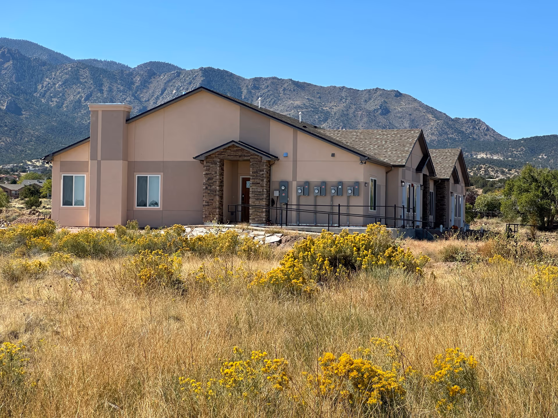 Exterior view of a single-story beige building with a stone entrance and multiple windows, surrounded by dry grass and yellow wildflowers, with mountains in the background under a clear blue sky.