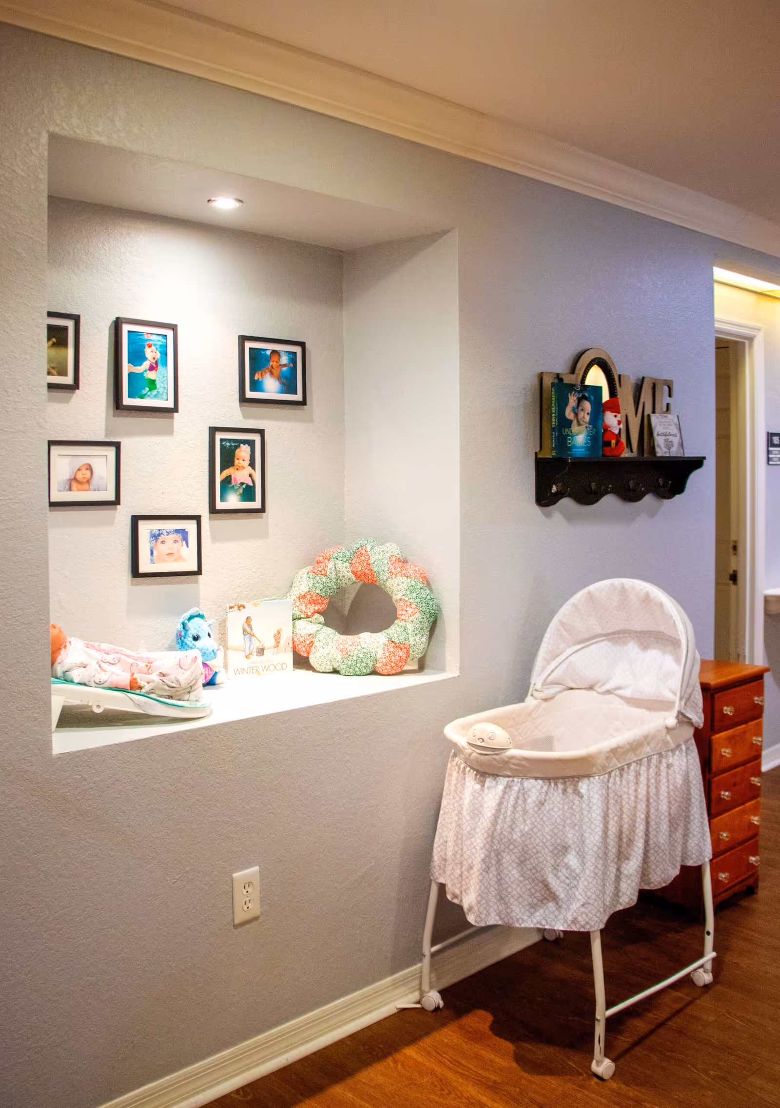 A baby bassinet sits beside a recessed wall niche filled with framed photos, toys, and decorative items in a hallway.