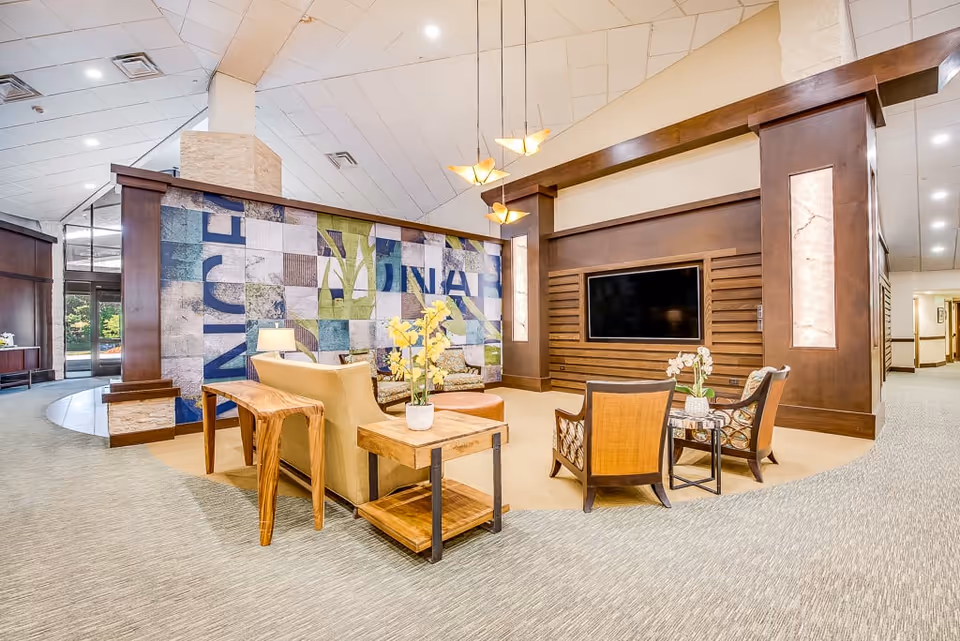 A bright and spacious lounge area in a senior living facility with comfortable seating including armchairs and a sofa arranged around a wooden coffee table and side tables with potted plants. The room features a large wall-mounted flat screen TV, decorative wooden paneling, modern pendant lights, and a colorful abstract wall mural. The carpeted floor and high ceiling add to the open and welcoming atmosphere.