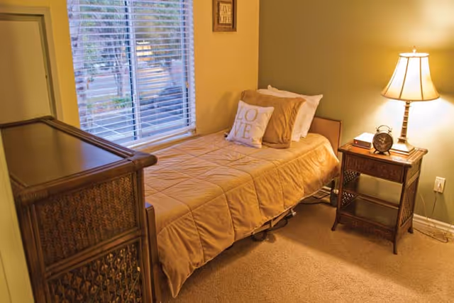 A cozy bedroom with a single bed covered in a beige quilt and pillows, including one with the word 'LOVE'. Next to the bed is a wooden nightstand with a lamp, an alarm clock, and a small book. A window with white blinds lets in natural light, and a wooden dresser is positioned near the foot of the bed.