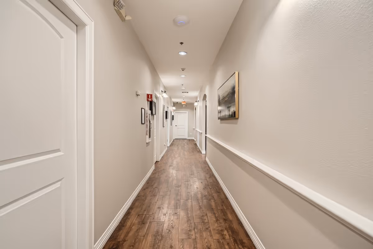 A long, narrow hallway with wooden flooring and beige walls. Several white doors line the left side, and a painting hangs on the right wall. The ceiling has recessed lighting and smoke detectors. A fire extinguisher and emergency signage are mounted on the left wall.