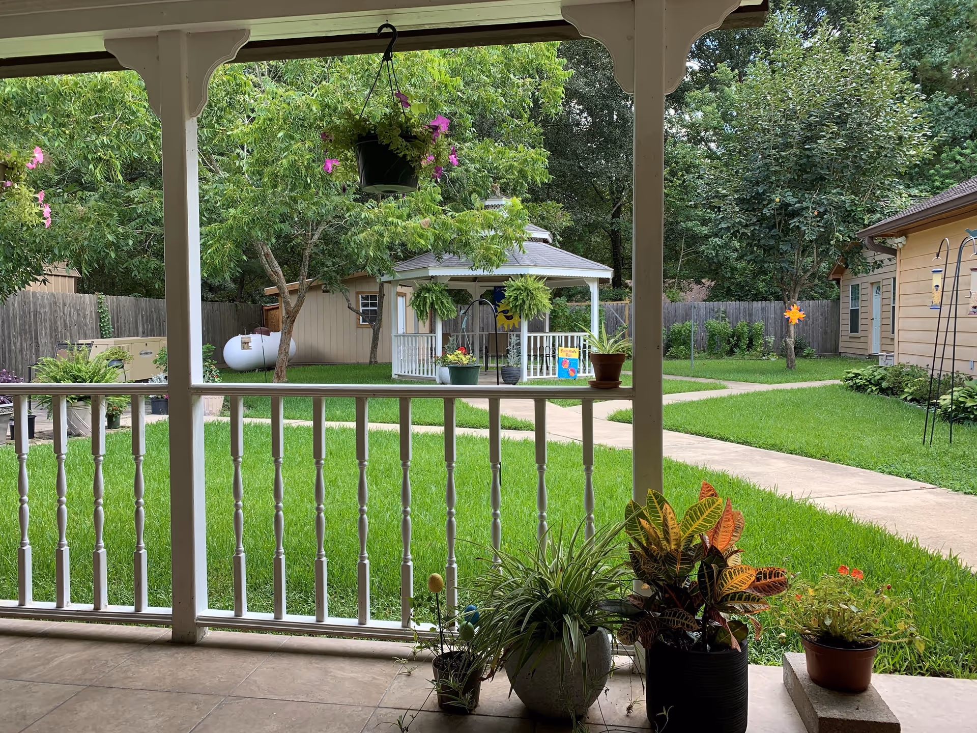 View from a covered porch overlooking a grassy backyard with potted plants, a gazebo, walkway, and surrounding trees and sheds.