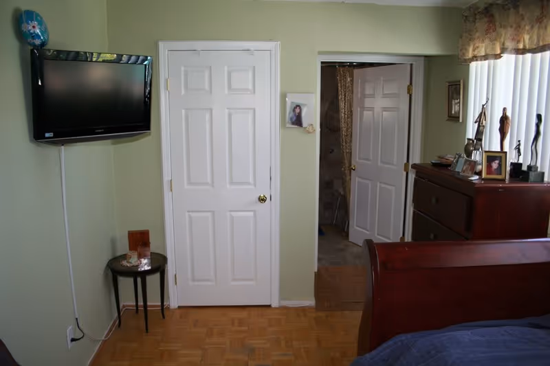 A bedroom with light green walls, a mounted flat-screen TV on the left wall, a small round table beneath the TV, a white door in the center, and an open door leading to a bathroom. On the right side, there is a wooden dresser with framed photos and decorative statues, next to a window with vertical blinds and a floral valance. The corner of a wooden bed frame with a blue bedspread is visible in the foreground.