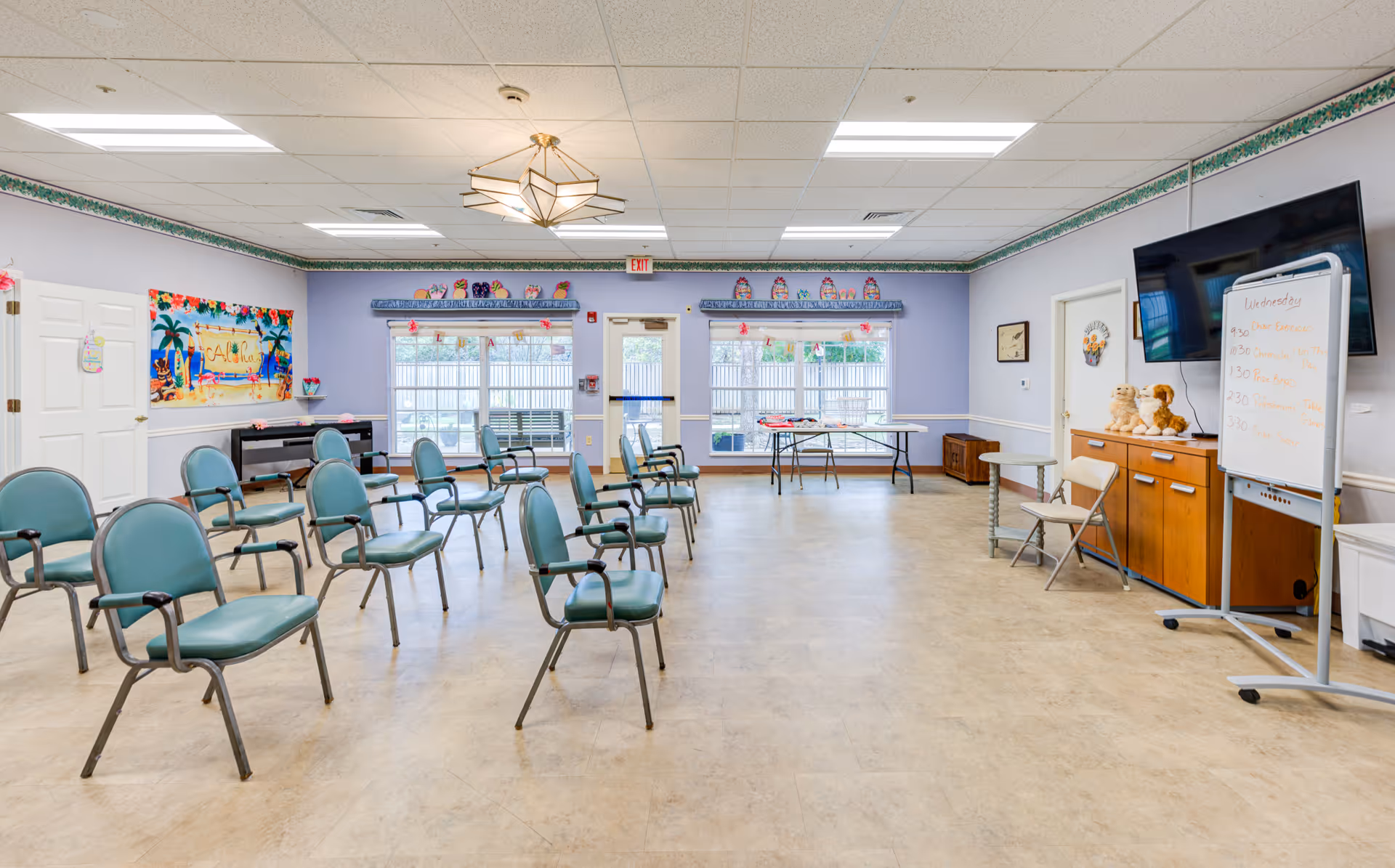 A spacious activity room with multiple green chairs arranged in rows facing a whiteboard and a large TV mounted on the wall. The room has light purple walls with a floral border near the ceiling, large windows and a door leading outside, a colorful 'Aloha' banner on one wall, and a table with various items near the windows. There are also two stuffed animal toys on a wooden cabinet and a small round table with a folding chair.