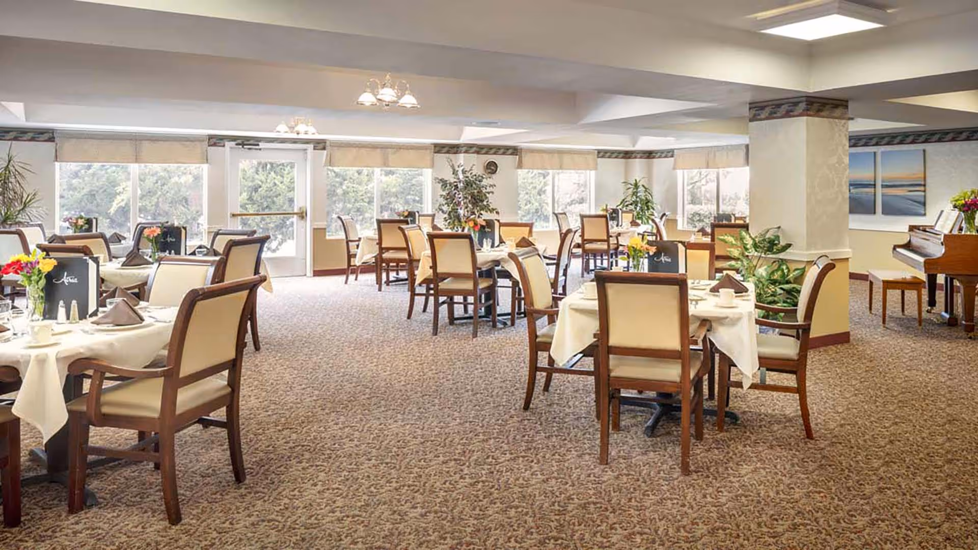 A bright and spacious dining room with multiple tables covered in white tablecloths, each set with napkins, cups, and small flower arrangements. The room has large windows letting in natural light, beige carpet, and several plants placed around. A piano is visible in the corner near a column.