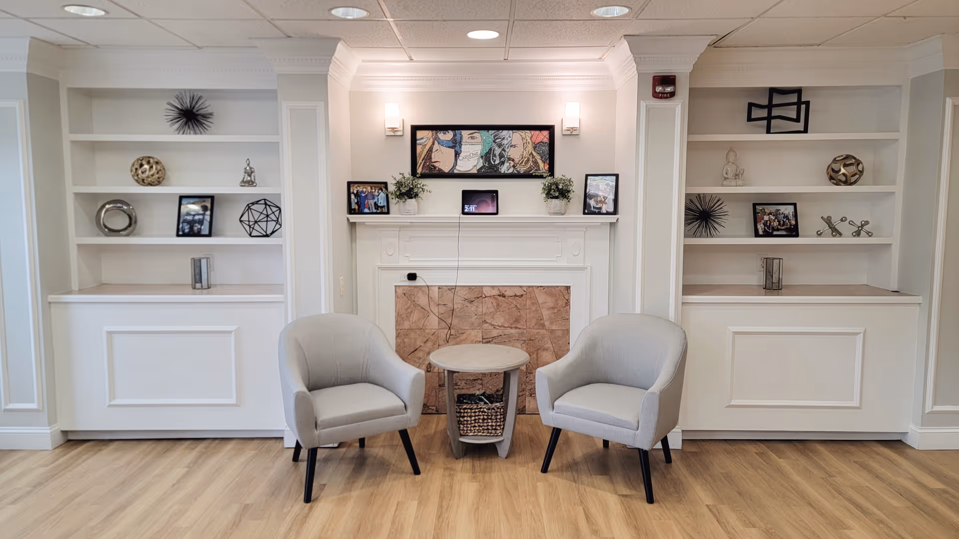 A cozy seating area in a senior living facility with two light gray armchairs positioned around a small round table. Behind the chairs is a white fireplace with a marble surround, decorated with framed pictures, plants, and a colorful artwork above. Built-in white shelves flank the fireplace, displaying decorative items such as sculptures, framed photos, and geometric objects. The room has light wood flooring and soft lighting from wall sconces.