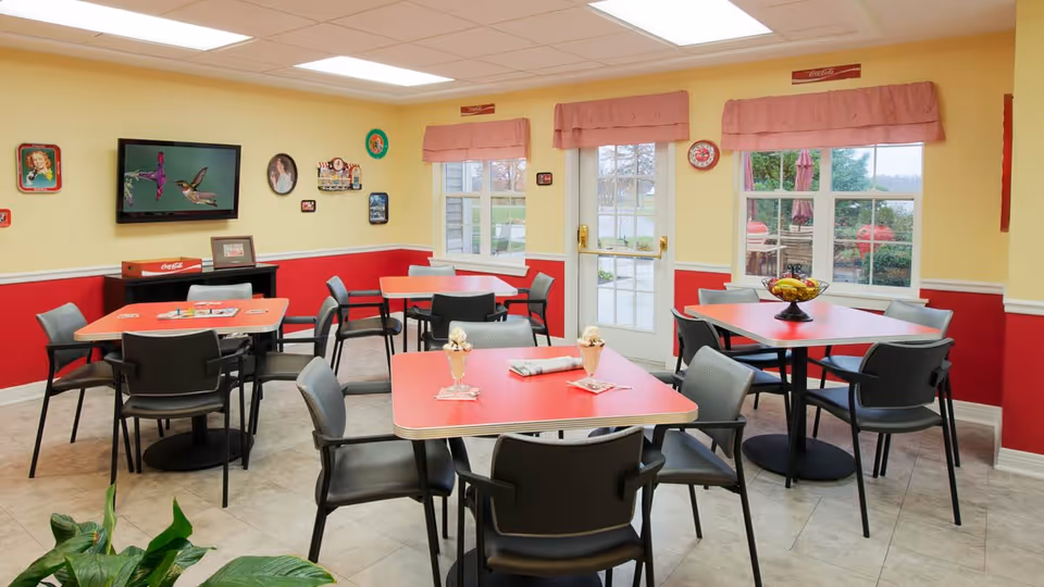 A bright dining room with red and yellow walls, featuring several red tables with black chairs arranged around them. The room has large windows with pink valances, a door leading outside, and a wall-mounted TV displaying an image of hummingbirds. Decorative plates and pictures adorn the walls, and a bowl of fruit and two ice cream sundaes are placed on the tables.