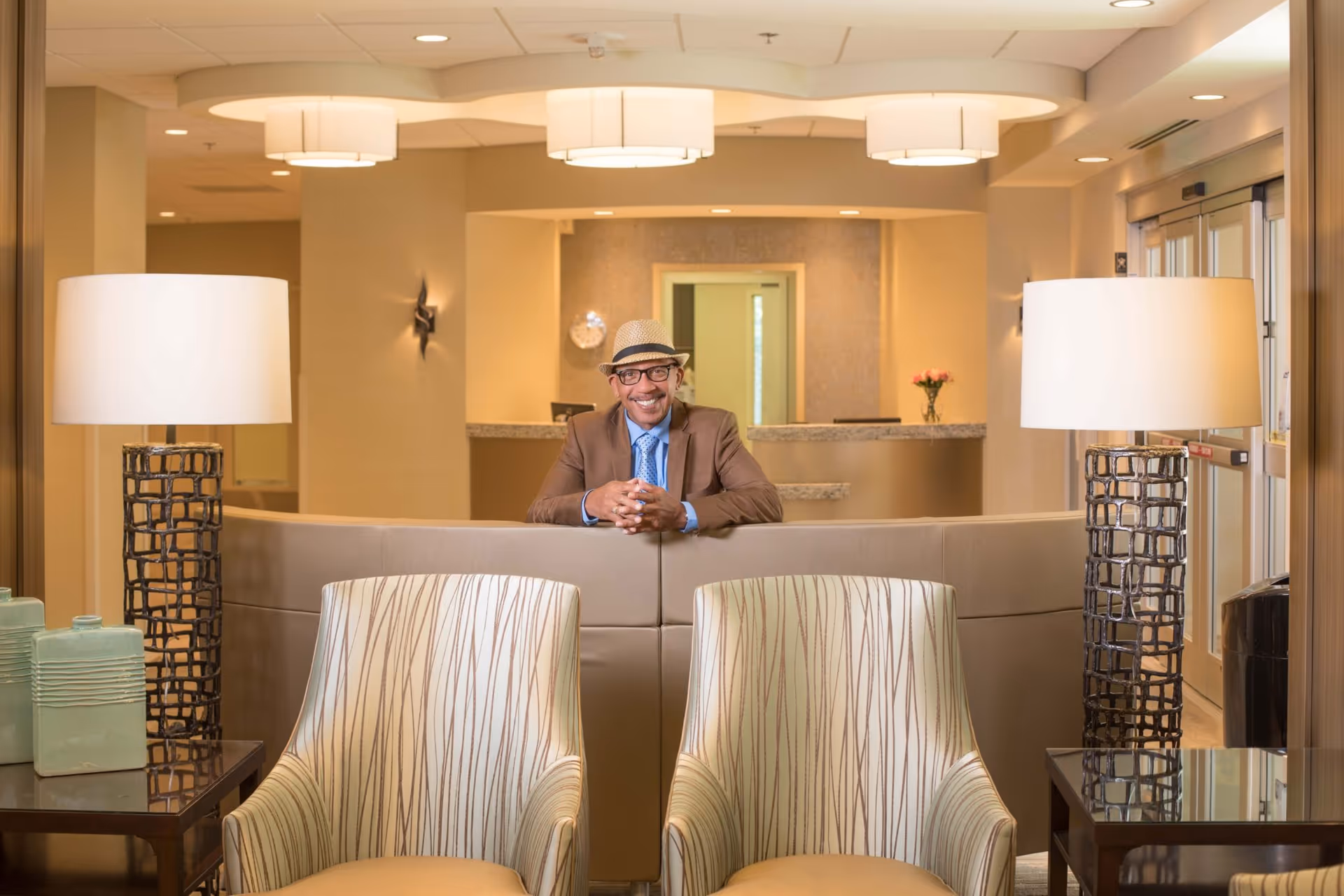 A man wearing a hat, glasses, a brown blazer, and a blue shirt with a tie is smiling and leaning on the back of a beige sofa in a well-lit reception or lounge area. The room features two patterned armchairs in the foreground, two modern table lamps with white shades on either side, and a reception desk with a vase of flowers in the background.