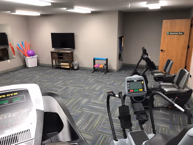 Interior view of a fitness room with exercise equipment including a treadmill and stationary bikes. The room has a patterned carpet, a wall-mounted TV, a mirror, and a basket with colorful exercise balls and foam noodles. A door labeled 'Exercise Rm' is visible on the right side.