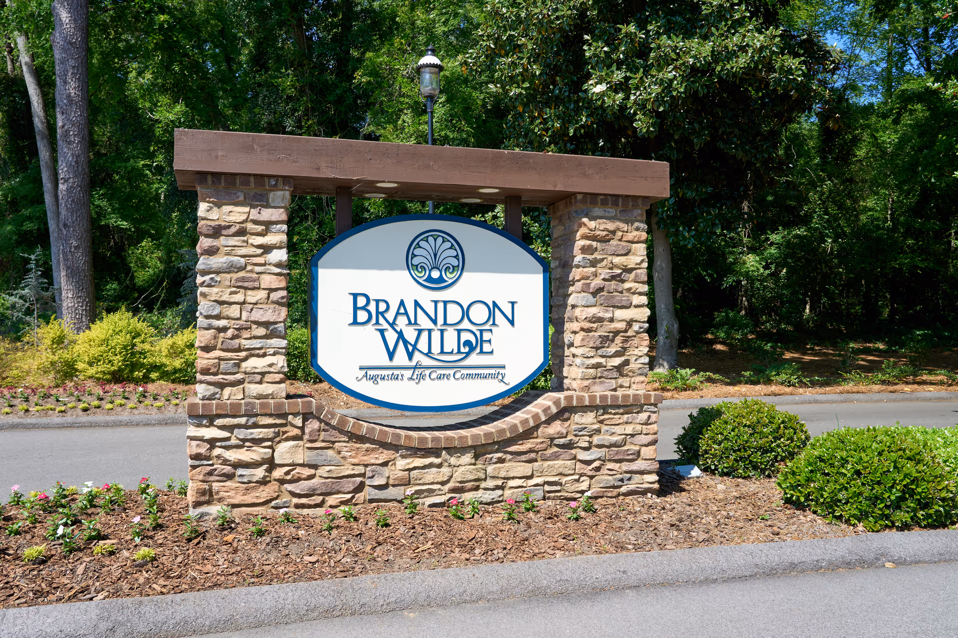 Stone and wood entrance sign for Brandon Wilde, Augusta's Life Care Community, surrounded by landscaping with bushes, flowers, and trees in the background.