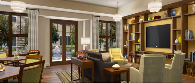 A bright communal lounge with sofas and armchairs facing a built-in shelving unit and TV, dining tables to the side, and glass doors opening to a garden patio.