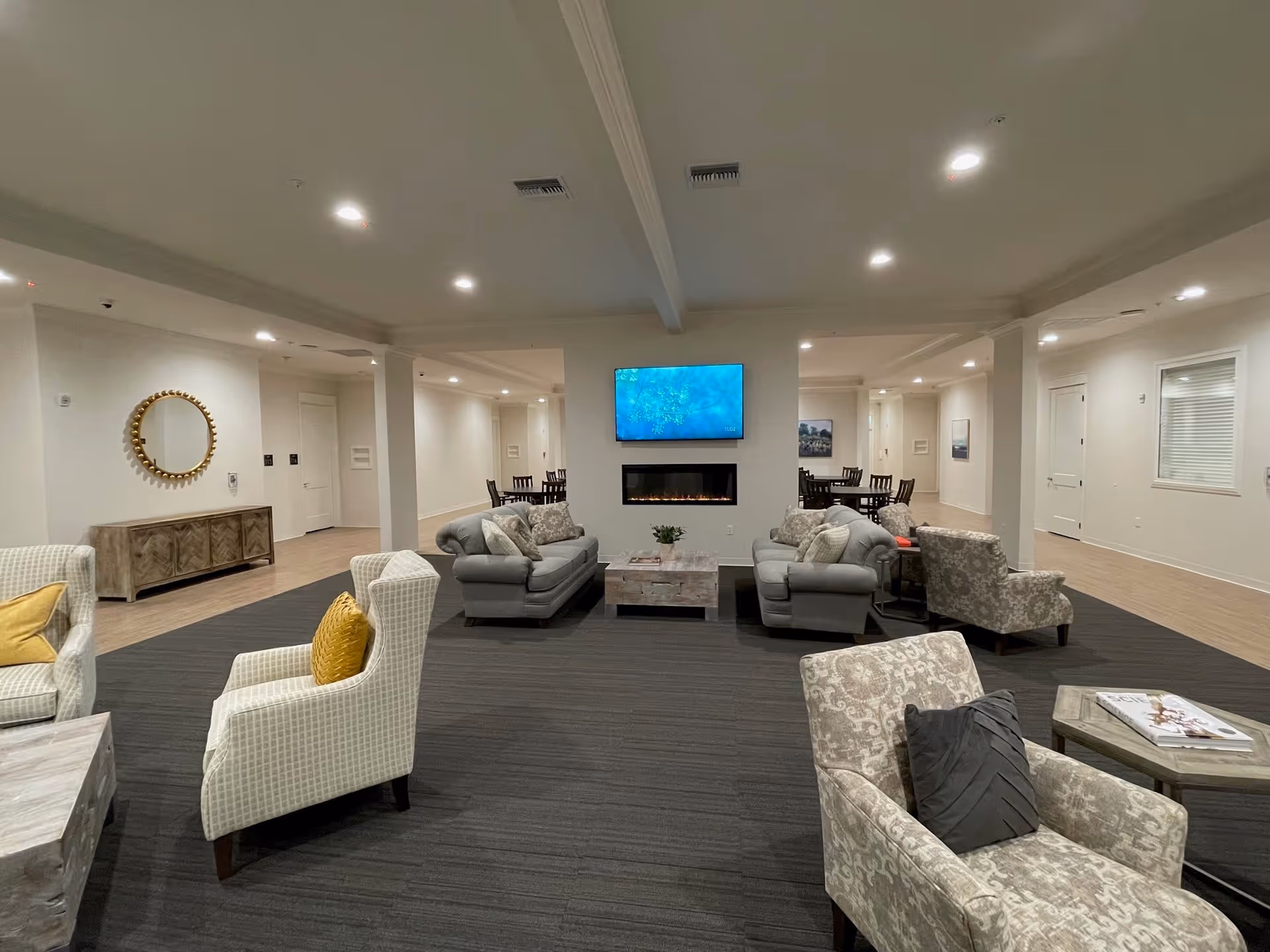 A spacious and well-lit common area in a senior living facility with multiple seating arrangements including sofas and armchairs with cushions. A modern electric fireplace is built into a central wall beneath a mounted flat-screen TV. In the background, there are dining tables and chairs along the walls. The room has a neutral color palette with light walls and dark carpeted flooring in the seating area, and wood flooring in the dining area.