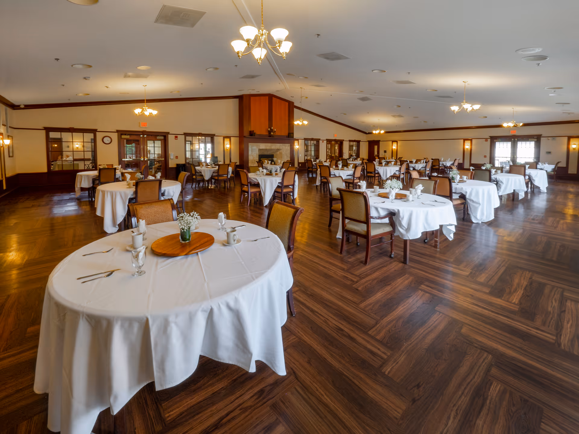 Large dining hall with round tables set with white tablecloths and chairs on a wood-patterned floor.