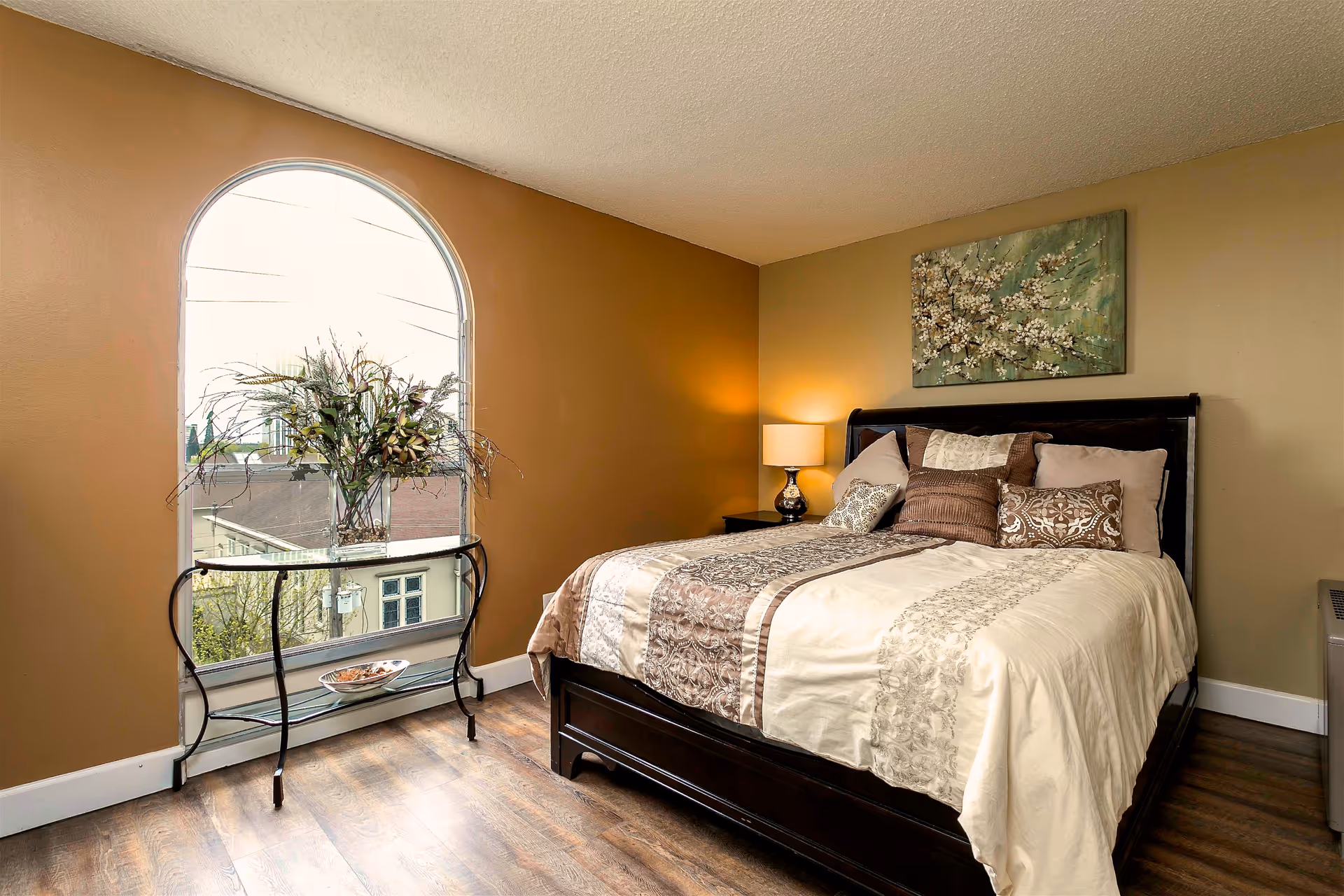 A neatly made bedroom with a dark wood bed, bedside lamp, decorative wall art, and an arched window with a glass console table holding a vase of flowers.