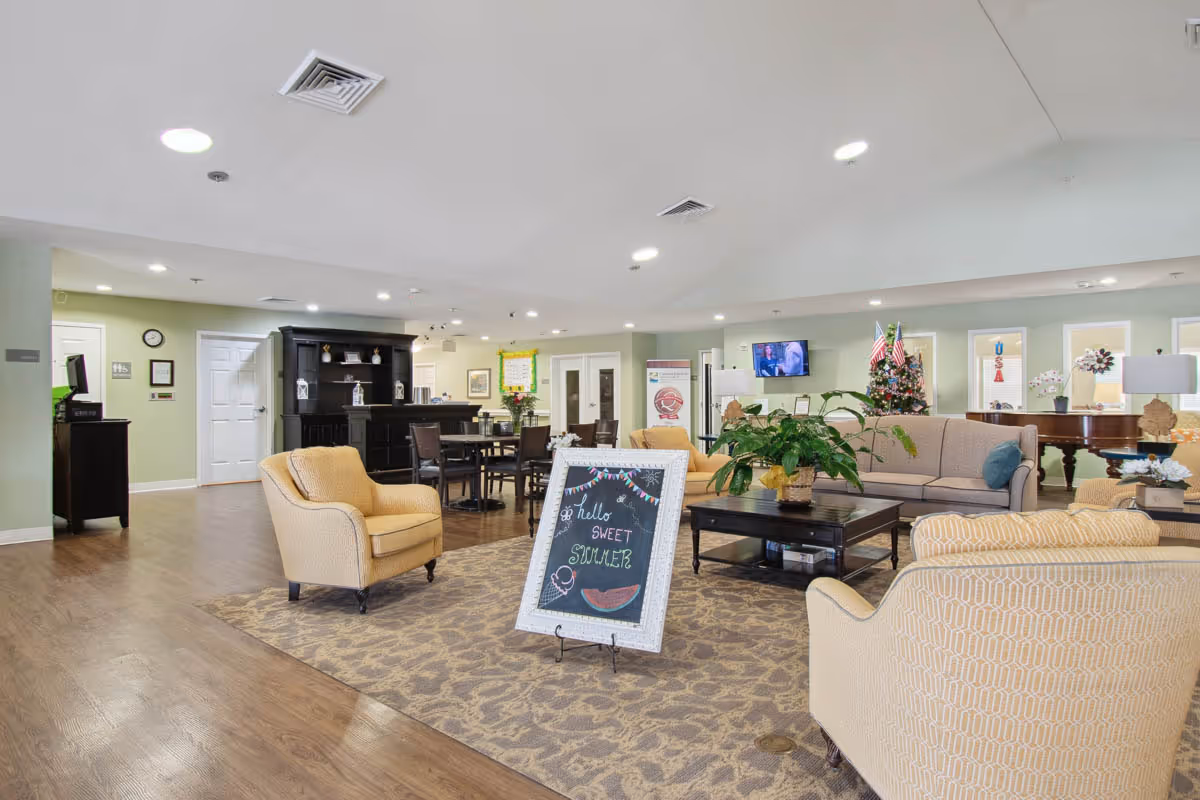 A spacious and well-lit senior living facility common area with beige armchairs, a beige sofa, a dark wooden coffee table with a plant, and a chalkboard sign that reads 'hello SWEET SUMMER'. In the background, there is a dining table with chairs, a black cabinet, a television mounted on the wall, and a decorated Christmas tree. The room has light green walls, wooden flooring, and ceiling lights.