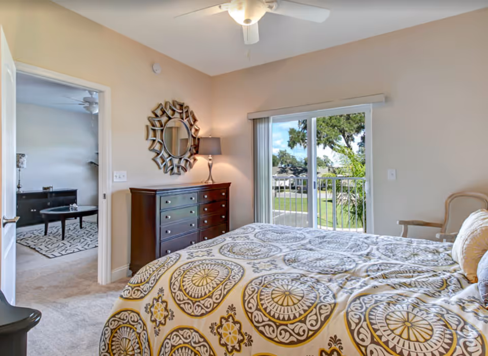 A bedroom with a patterned bedspread in shades of yellow, gray, and white. The room features a dark wooden dresser with a decorative round mirror above it and a table lamp. There is a sliding glass door leading to a balcony with a view of green trees and a lawn outside. An open door reveals a glimpse of an adjacent room with a coffee table and a patterned rug.