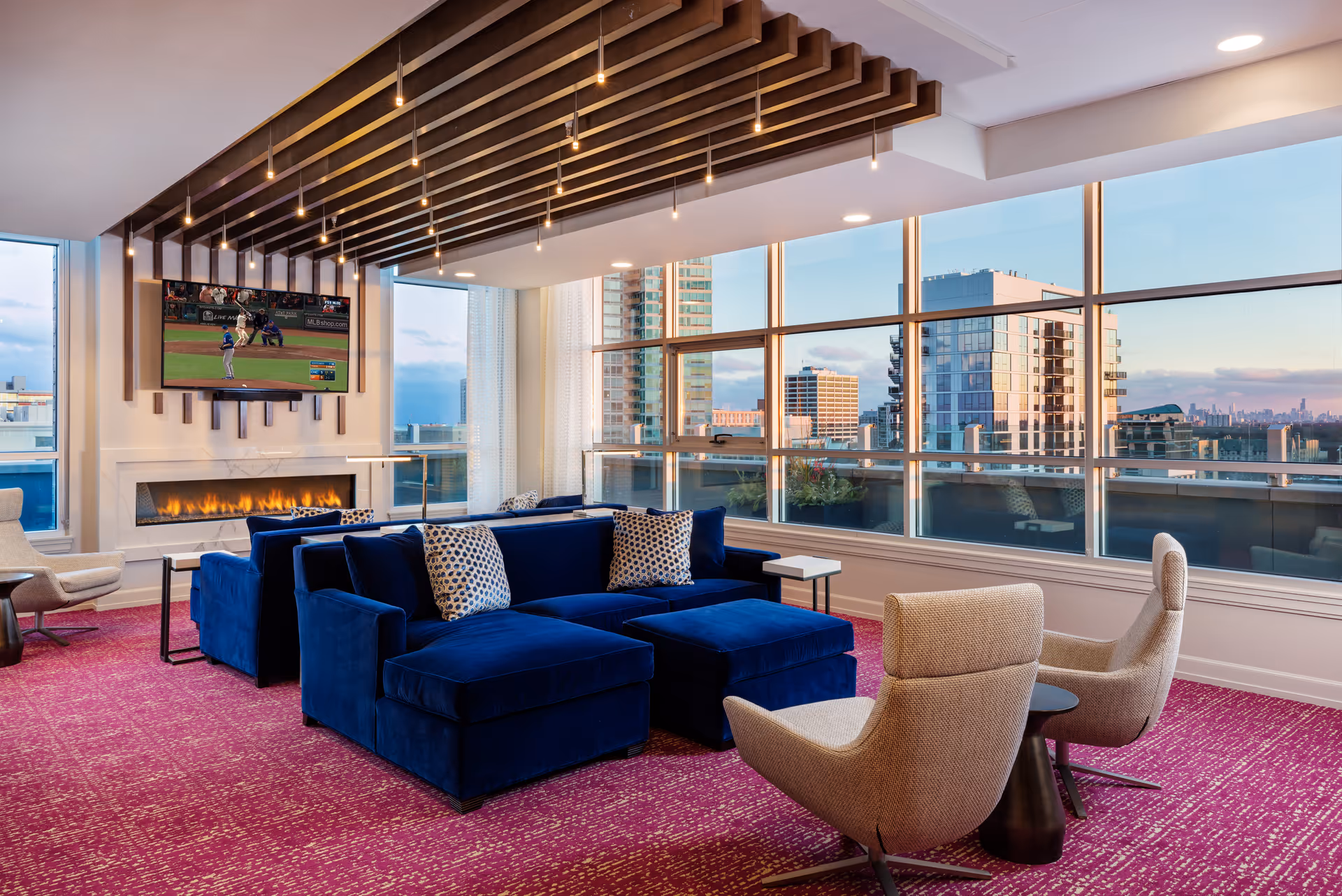 Modern living room with large windows offering a cityscape view, featuring a blue sectional sofa with patterned pillows, two beige armchairs, a fireplace beneath a wall-mounted TV showing a baseball game, and a ceiling with wooden slats and hanging lights. The floor is covered with a red patterned carpet.